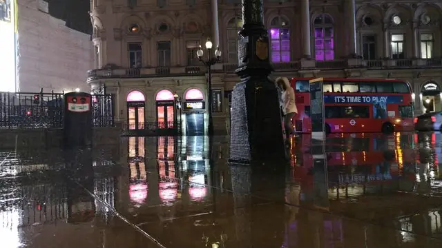 Double-decker bus in rainy Central London at night with street reflections.